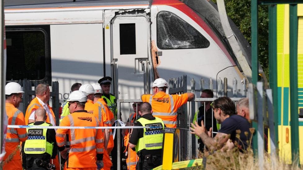 A large crowd of police and railway workers attending a damaged white train carriage.