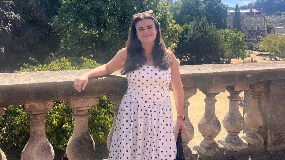 Ella Kipling, a young woman with long dark hair, smiles at the camera. She is wearing a long, white dress with black polkadots and is standing in front of a large park with old buildings in the background.