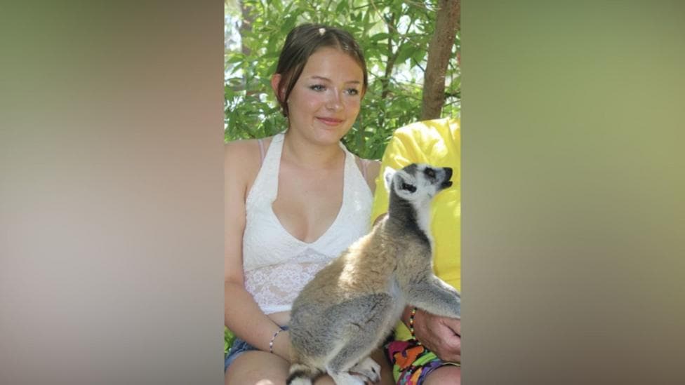 Lillie Nichols has tied back brown hair, a white vest, denim shorts with a ring-tailed Lemur on her knee.