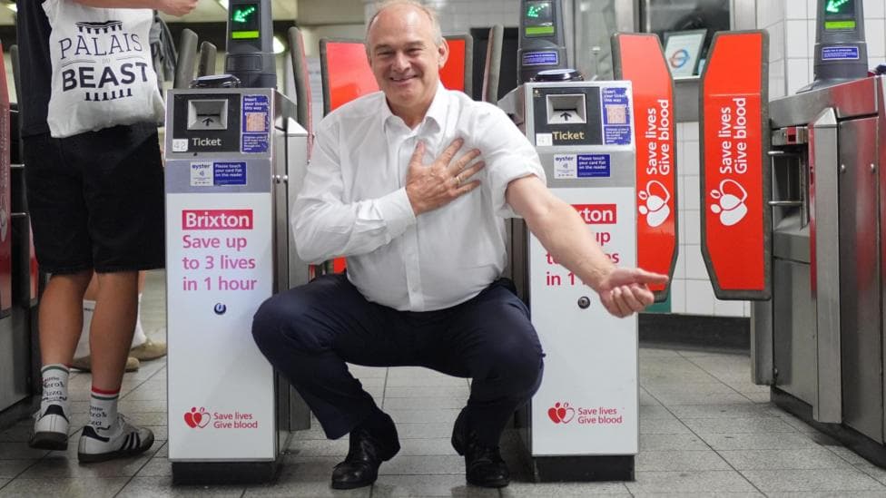 Ed Davey crouches in front of tube barriers, which are advertising the nearby blood donation centre. The adverts read: "Brixton: Save up to 3 lives in 1 hour." Ed Davey is wearing dark trousers and a white shirt with one sleeve rolled up as if about to donate blood.