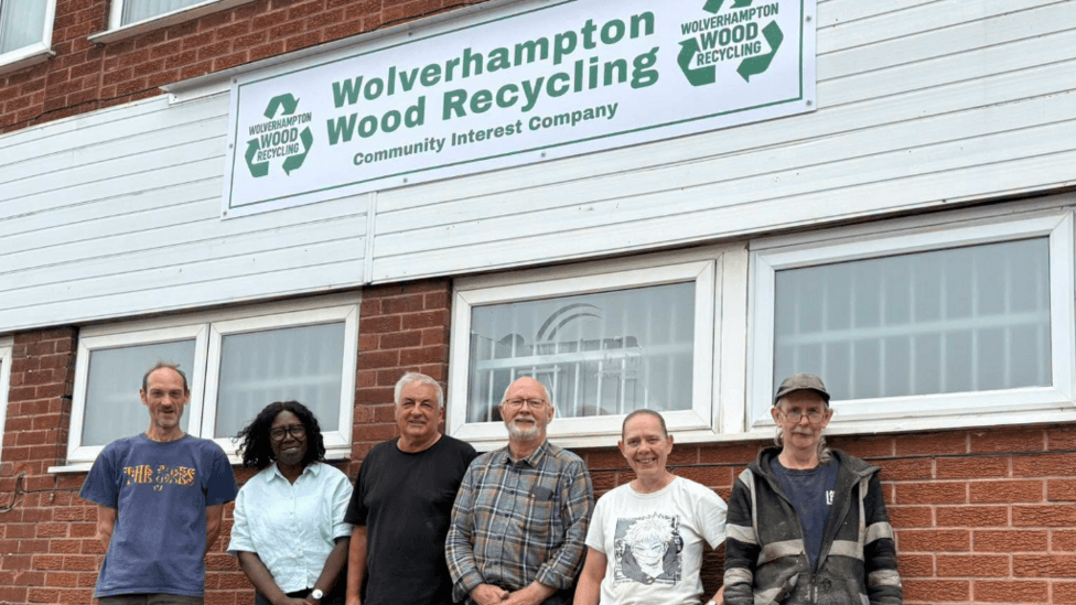 Six men and women stand outside the new premises. They stand below a sign of the recycling group. They are leaning against the building and are smiling at the camera.