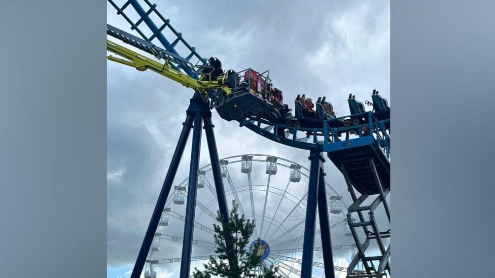 People in carriages on a rollercoaster. They are high off the ground. The sky is blue and there are white clouds. A white Ferris wheel is also featured.