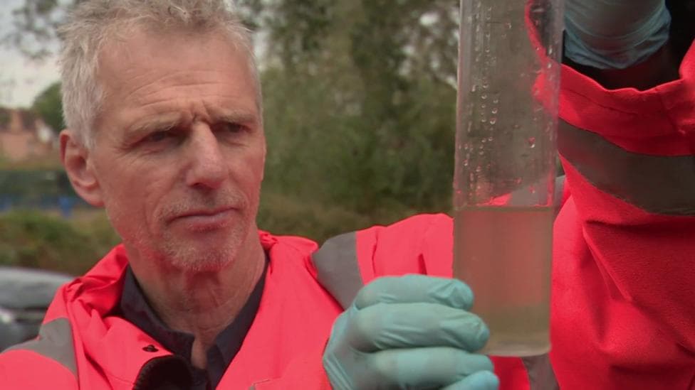 Man in a pink hi-viz coat and rubber gloves holding a jar of murky water while he stares at it in a concerned manner.