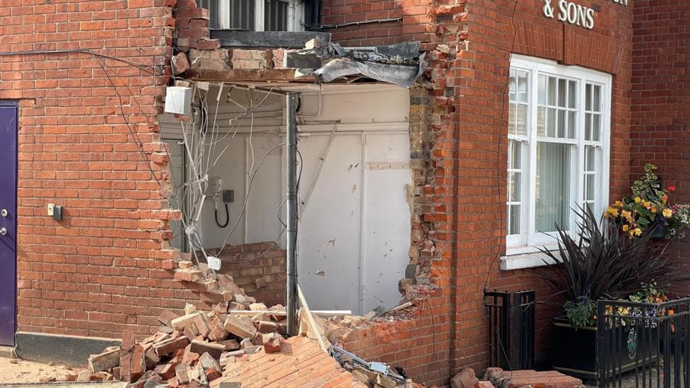 A massive hole in a wall with rubble strewn on the floor. There is a white wooden-framed window to the right, with various plants in front.