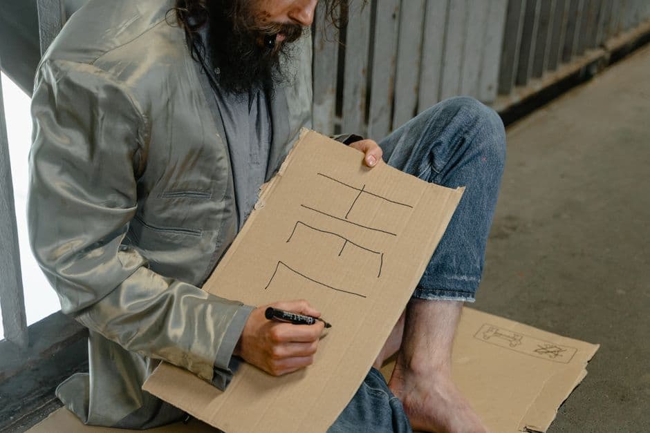 A homeless man writing 'Help' on a cardboard sign in an urban environment, reflecting poverty.
