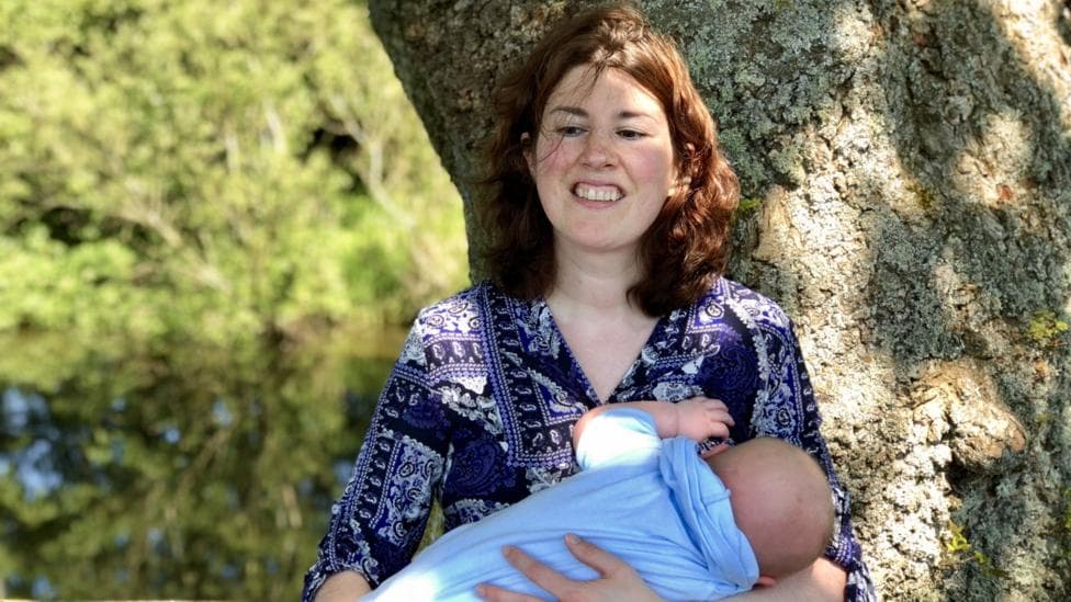 BBC reporter Emma Tracey is pictured holding a baby wrapped in a light blue sling while standing beside a large tree and wooden fence. Sunlight filters through surrounding greenery, with water and blurred trees visible in the background.