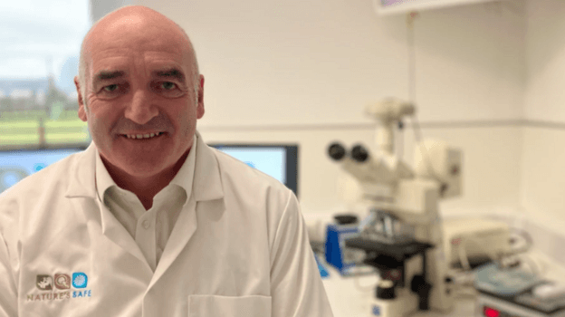 Photograph of Matson who is wearing a white shirt and a white coat with "Nature's Safe" embroidered on it. He's pictured inside a lab, with a large microscope on a desk behind him. The room has white walls and a few screens on the desk.