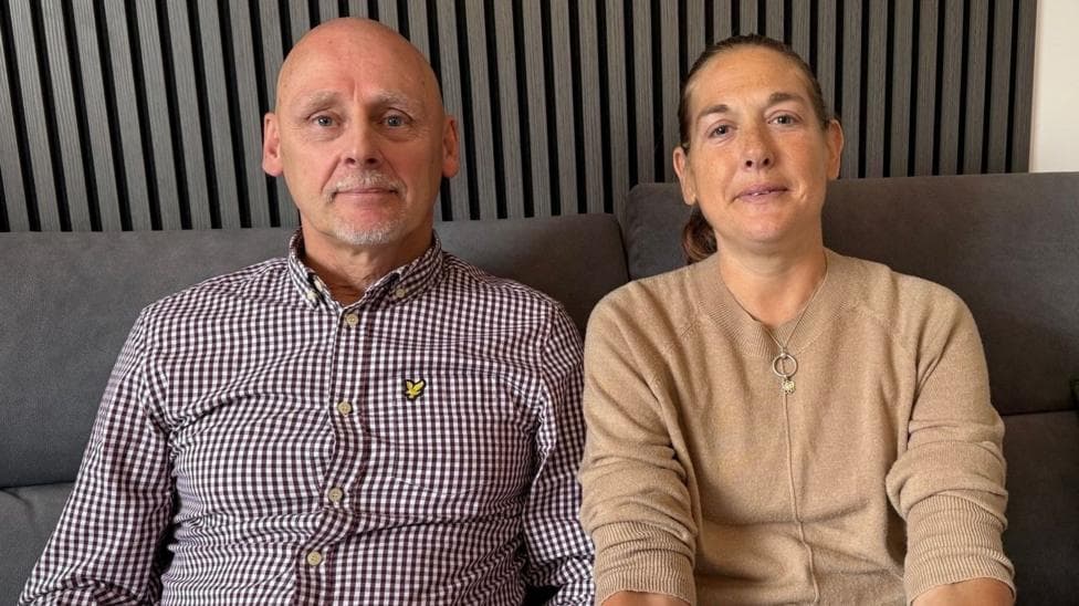 Gary is bald and wears a burgundy and white checked shirt and sit nexts to Karen who has her hair in a ponytail is wearing a camel coloured jumper.