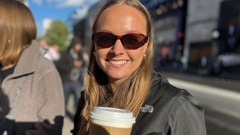 A portrait shot of Emily staring at the camera. She is wearing dark glasses and a black jacket, and has light brown hair. She us holding a cup of coffee in front of her.
