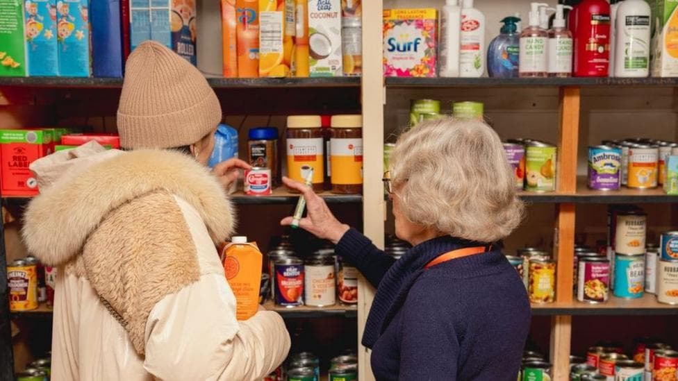 There are two people stood in front of shelves of food cupboard items. The taller person on the left is wearing a large beige coat and a brown beanie. The shorter woman on the left has short grey hair, is wearing a purple top and pointing to a tin on the shelf.
