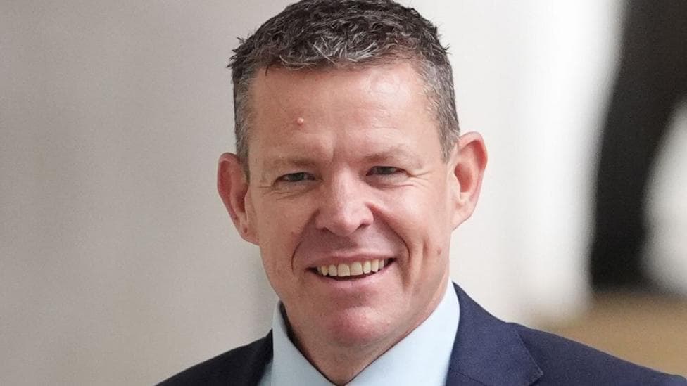 A close up of Rhun ap Iorwerth's face - he is wearing a dark blue suit, light blue shirt and is stood in front of a background that is out of focus.