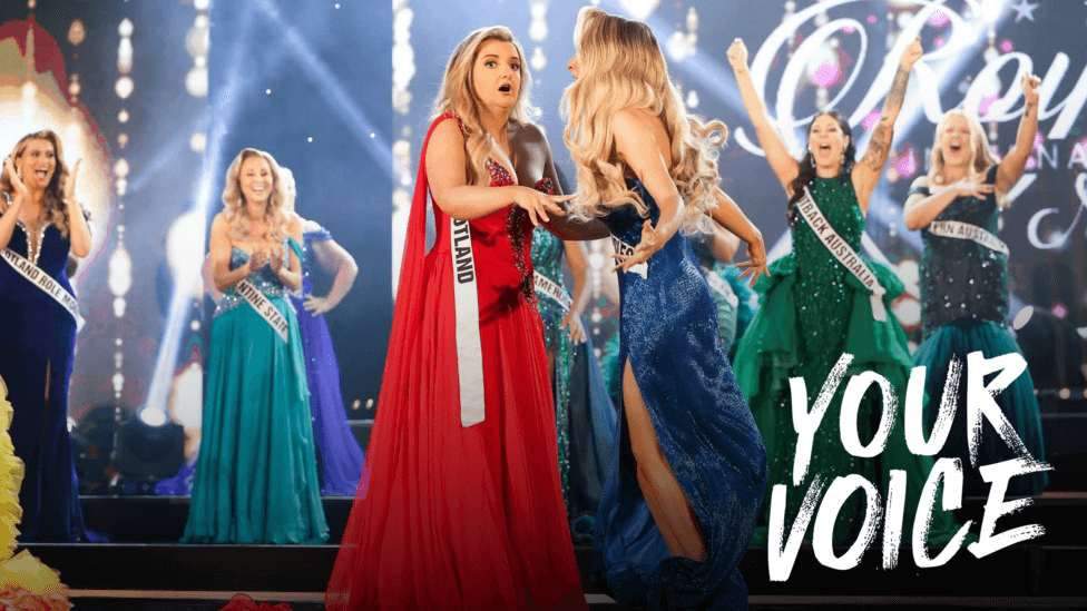 A brightly lit beauty pageant stage during the announcement of a winner. At centre stage, a contestant in a flowing red gown and a sash reading “Scotland” is congratulated by another contestant in a sparkling blue gown.
Several other sash-wearing contestants in colourful evening dresses stand behind them on tiered steps.