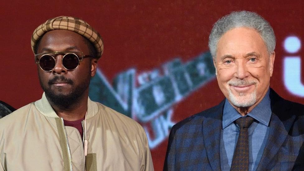Will.I.Am stands next to Tom Jones, Will.I.Am is wearing a checked hat, sunglasses and a beige jacket. Tom Jones is wearing a blue shirt and checked jacket with a checked tie.