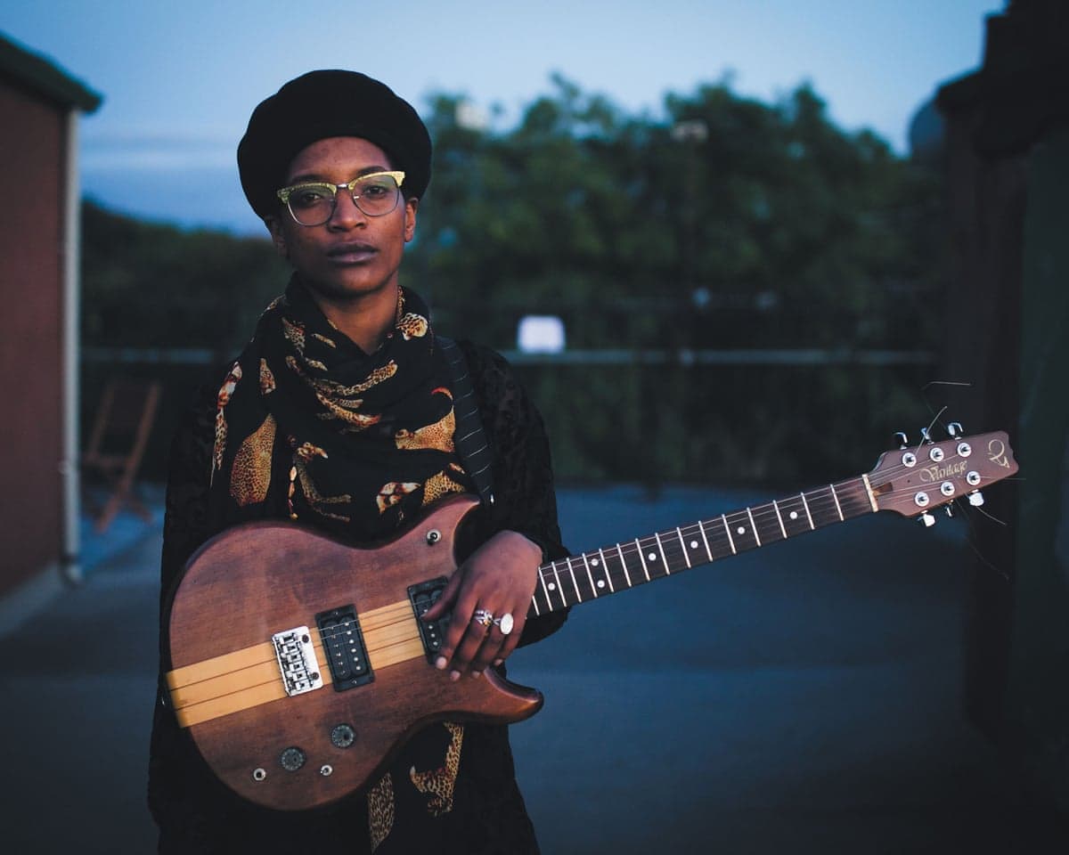 Beldina Odenyo, in glasses and beret, holds an electric guitar, looking at the camera