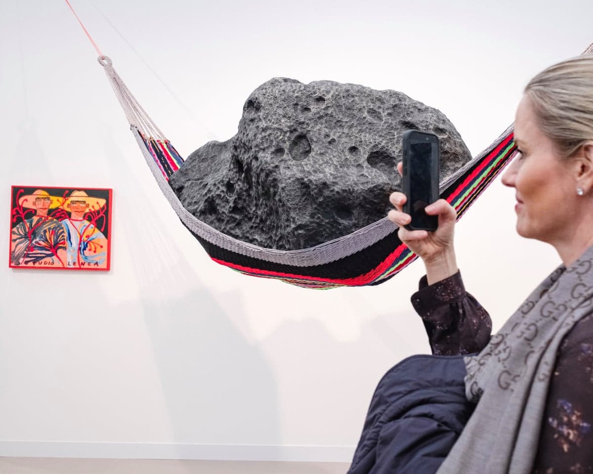 A visitor photographs a large rock sculpture suspended in a woven hammock on the Carl Freedman stand at Frieze London