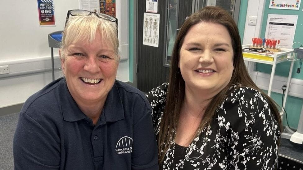 Karen Pyle, left, and Jessica Beni, right, are sitting down in a classroom. Karen is blonde and is wearing a navy polo neck top with Newcastle College emblem on the right hand side. Jessica has long dark hair and is wearing black dress with a white floral pattern on it. Behind them are grey double doors with chrome handles and narrow glass windows, there is a table with stationery and scissors on and some posters on the wall.