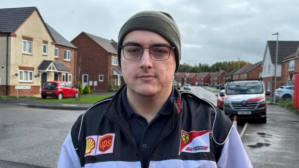 There is a man in a green beanie, black glasses and a black and white jacket. On his jacket, there are three logos, the Shell petrol logo, the UPS logo and the Scuderia Ferrari Formula 1 logo. He is stood on a residental street with new-build homes to his left and right.