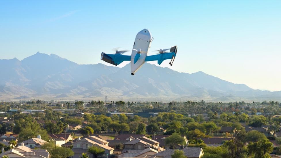 An Amazon drone flies over an US city. In the background are mountains and a blue sky.