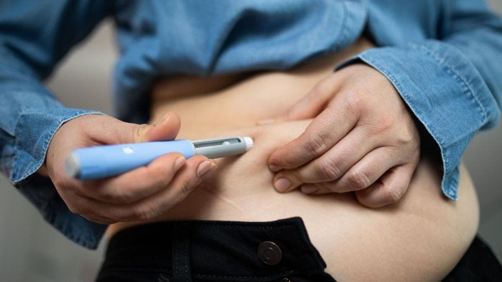 A person injecting a medication into their stomach - they are wearing a denim shirt and black trousers