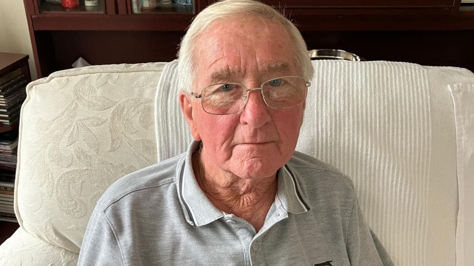 A photo of an older man - grey hair and glasses. He's sitting on a cream coloured sofa and is wearing a grey polo top