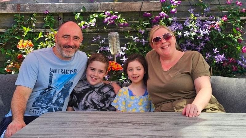A family of four sitting at a table in a garden, with various flowers behind them. The dad, Scott, is bald and wearing a blue T-shirt with San Francisco written on it. Mum Laura is wearing sunglasses and a green dress. Sitting in-between their parents are son Aidan and daughter Grace. All are smiling for the camera.